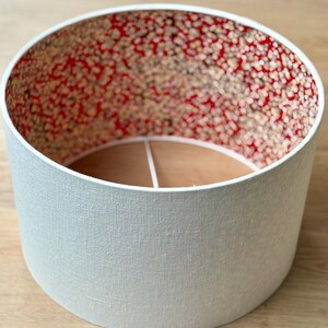 May include: A cylindrical lampshade with a pale grey exterior and a patterned interior. The interior has a red background with small, speckled designs in shades of brown and cream. The lampshade is designed to fit over a light fitting.