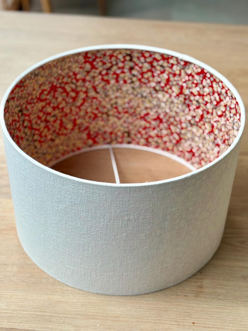 May include: A cylindrical lampshade with a pale grey exterior and a patterned interior. The interior has a red background with small, speckled designs in shades of brown and cream. The lampshade is designed to fit over a light fitting.