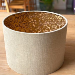 May include: A cylindrical lampshade with a neutral beige exterior. The interior features a detailed floral pattern in shades of gold and brown. The lampshade is sitting on a wooden surface.