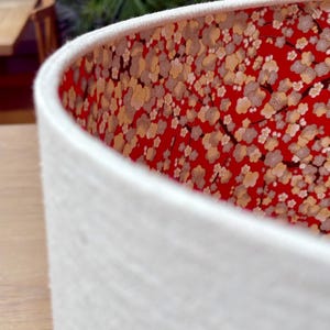 May include: A white lampshade with a textured exterior. The interior features a vibrant red fabric adorned with a floral pattern in cream and gold. The lampshade's design adds a touch of elegance to any room.