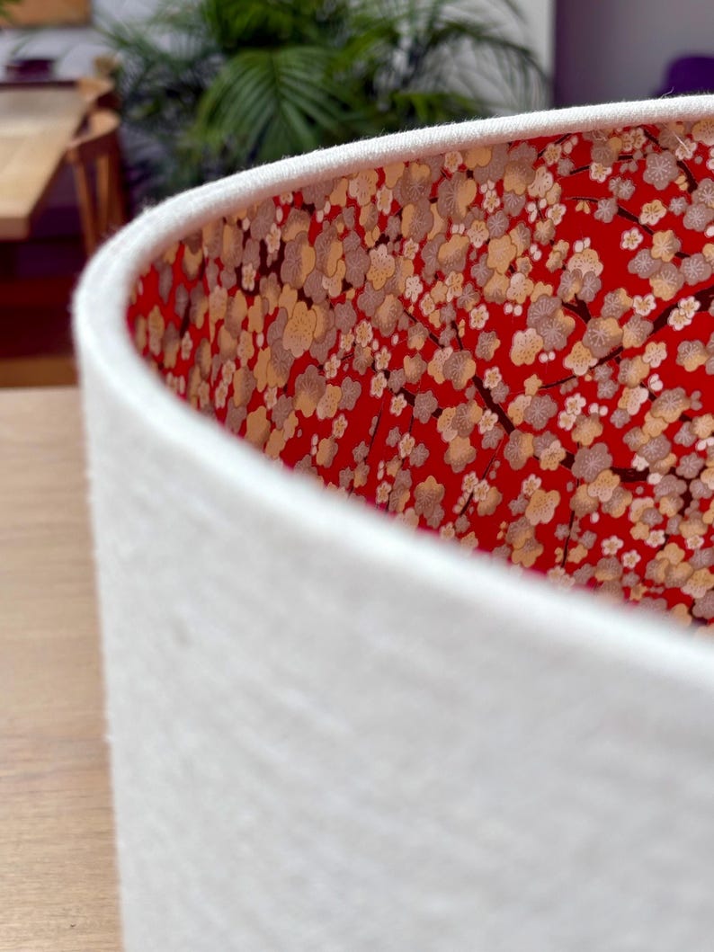 May include: A white lampshade with a textured exterior. The interior features a vibrant red fabric adorned with a floral pattern in cream and gold. The lampshade's design adds a touch of elegance to any room.