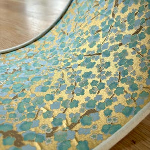 May include: Close-up of a decorative item with a gold background and a pattern of blue-green flowers and brown branches. The item has a white border and is curved, revealing the intricate floral design.