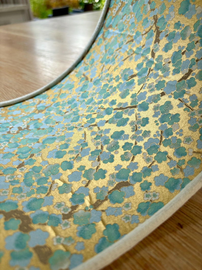 Op de afbeelding: Close-up van een decoratief item met een gouden achtergrond en een patroon van blauwgroene bloemen en bruine takken. Het item heeft een witte rand en is gebogen, waardoor het bloemmotief zichtbaar wordt.