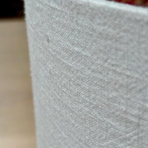 May include: Close-up of a cylindrical, off-white fabric container. The fabric has a textured, woven appearance. The interior reveals a glimpse of a patterned, reddish-brown material. The container's edge is visible, showcasing the fabric's thickness and texture.