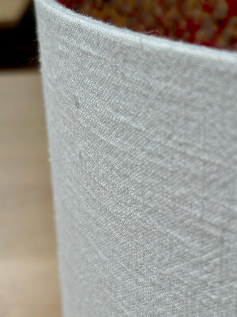 May include: Close-up of a cylindrical, off-white fabric container. The fabric has a textured, woven appearance. The interior reveals a glimpse of a patterned, reddish-brown material. The container's edge is visible, showcasing the fabric's thickness and texture.