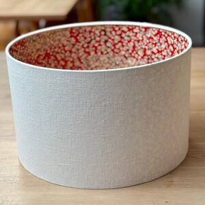 May include: A cylindrical lampshade with a textured white exterior and a red interior patterned with small, gold-coloured dots. The lampshade is sitting on a wooden surface.