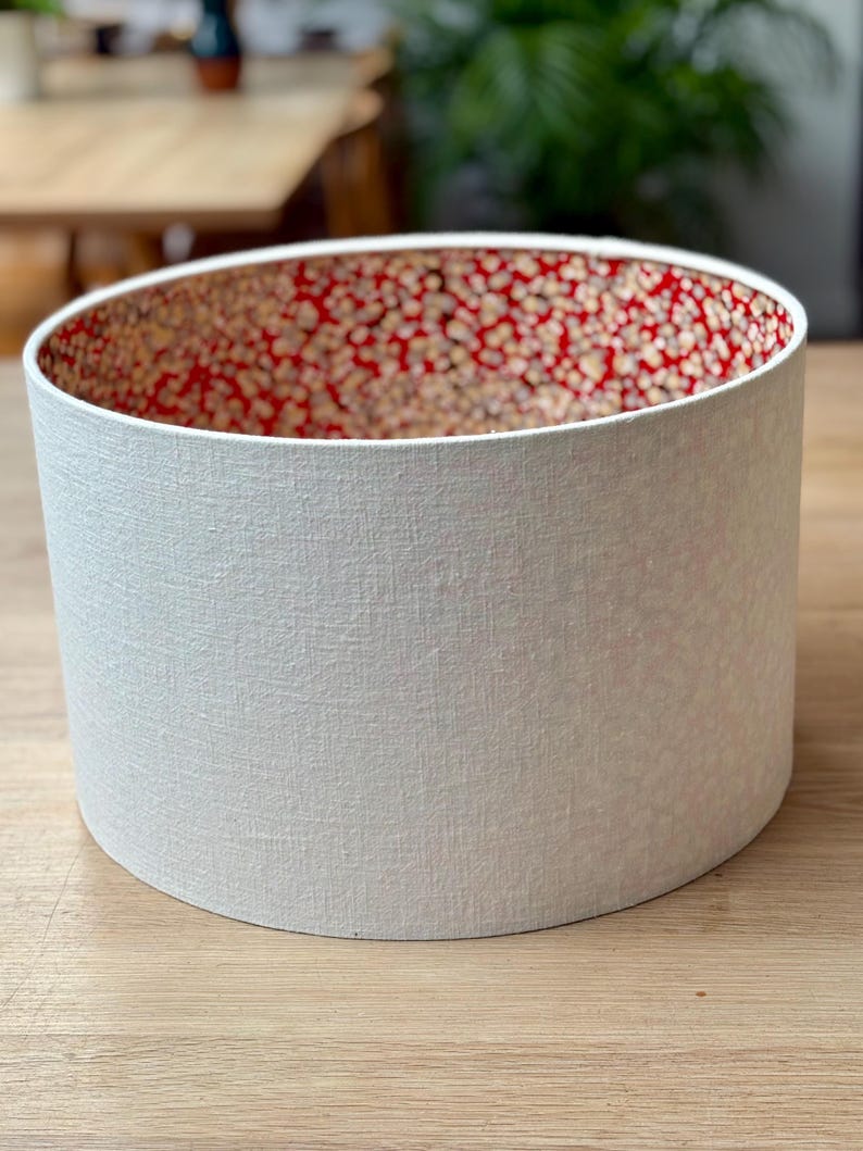 May include: A cylindrical lampshade with a textured white exterior and a red interior patterned with small, gold-coloured dots. The lampshade is sitting on a wooden surface.