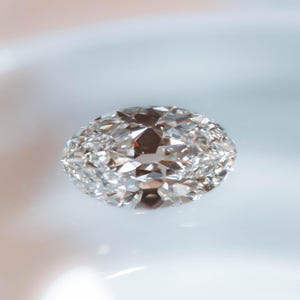 May include: A brilliant-cut, oval-shaped diamond. The gemstone is clear and colorless, with numerous facets that reflect light, creating a sparkling effect. The diamond is set against a soft, blurred background.