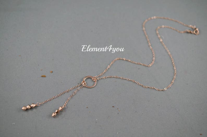 May include: A rose gold necklace with a circle pendant and two dangling strands of beads. The necklace is on a gray background.