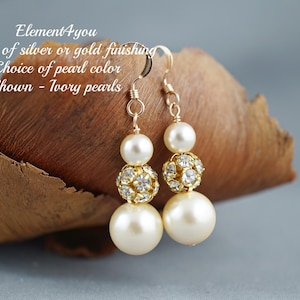 May include: A pair of ivory pearl and rhinestone dangle earrings. The earrings are made with gold-tone metal and feature a cluster of rhinestones at the bottom. The earrings are shown on a brown wooden surface.