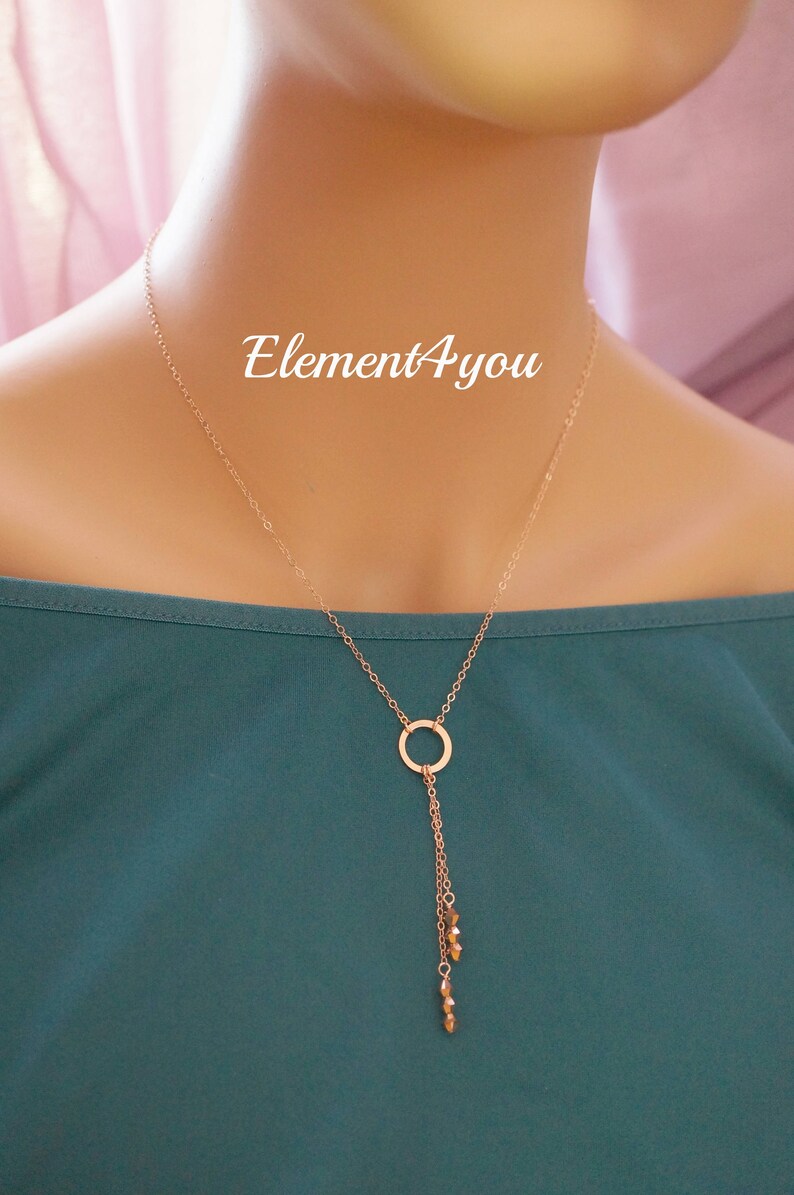 May include: A rose gold necklace with a circle pendant and a dangling chain with small gold beads. The necklace is on a mannequin wearing a teal shirt. The text "Element4you" is visible on the shirt.