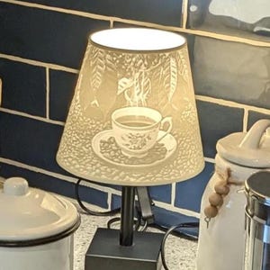 May include: A kitchen counter scene with a coffee-themed display. It includes a small lamp with a coffee cup design, a French press, and canisters labeled "COFFEE" and "SUGA". A two-tiered wooden tray holds a creamer, a small cup, and potted plants.