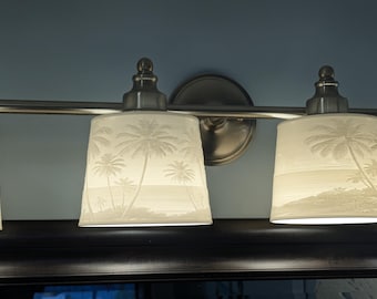 May include: A multi-light vanity fixture featuring a brushed nickel finish and cylindrical white glass shades. Each shade is decorated with a subtle, embossed tropical scene depicting palm trees along a shoreline, illuminated from within.