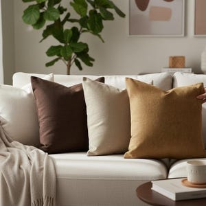 Op de afbeelding: Een verzameling sierkussens in wit, bruin en beige op een witte bank. Een neutrale deken is over de arm van de bank gedrapeerd. Een hand past een van de kussens aan. Een koffietafel met een mok.