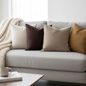 Op de afbeelding: Een lichtgrijze bank met vier decoratieve sierkussens in cr&egrave;me, bruin, beige en mosterdgeel. Een cr&egrave;mekleurige gebreide deken is over de arm gedrapeerd. Een salontafel met een boek en een mok staat voor de bank.