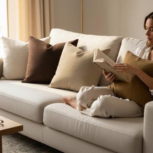 Op de afbeelding: Een cr&egrave;mekleurige bank met verschillende sierkussens in bruin- en beigetinten. Een persoon leest een boek op de bank. Een houten salontafel met een mok en een bril staat voor de bank.