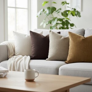 Op de afbeelding: Een lichtgrijze bank met vier decoratieve kussens in cr&egrave;me, bruin, beige en goud. Een cr&egrave;mekleurige deken is over de bank gedrapeerd. Een houten salontafel bevat een mok en boeken. Een kamerplant is zichtbaar op de achtergrond.