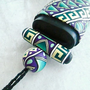 Puede incluir: Primer plano de un collar colorido y estampado con un cordón negro. El collar presenta una cuenta redonda, una cuenta cilíndrica y una cuenta cilíndrica más grande con una tapa negra. Las cuentas están decoradas con patrones geométricos en tonos verdes, morados y blancos.