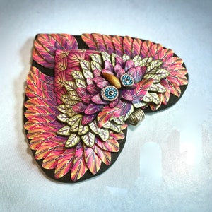 May include: A colourful owl sculpture made of polymer clay. The owl has pink, yellow, and white feathers and blue eyes. The sculpture is shaped like a heart.