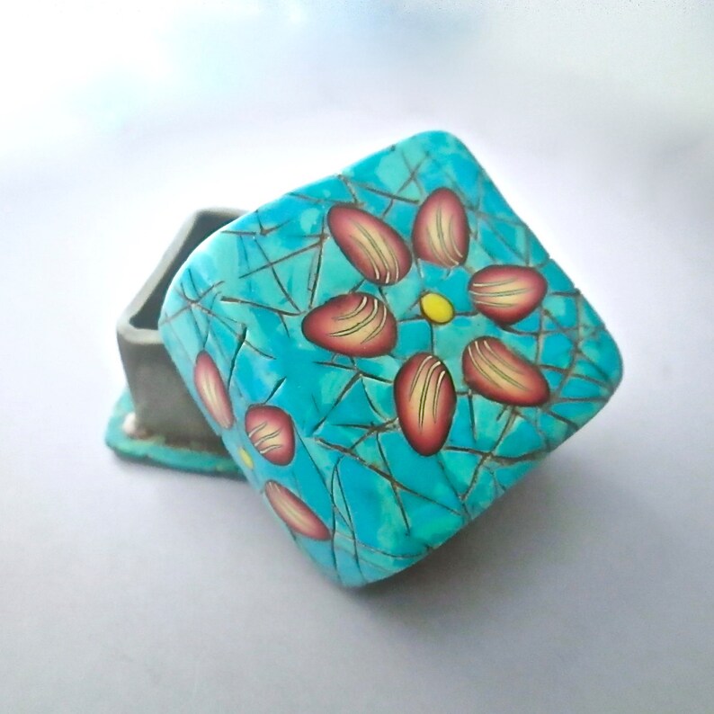 May include: A small, square, turquoise box with a red and yellow flower design. The box is made of polymer clay and has a crackled finish.