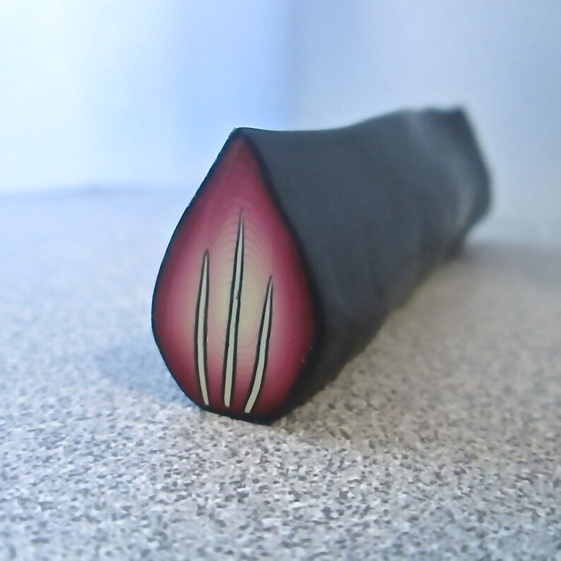 May include: A close-up of a polymer clay cane with a red and white design. The cane is cut in a teardrop shape, revealing a cross-section of the design.
