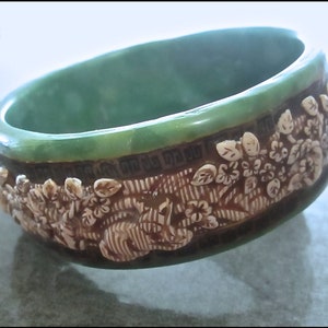 Pode incluir: Um bracelete de plástico verde com um design floral branco. O bracelete é decorado com um padrão floral em relevo e uma borda geométrica.