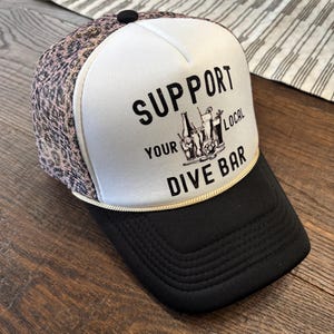 May include: A trucker hat with a black brim and a white front panel featuring the text "SUPPORT YOUR LOCAL DIVE BAR" in black. The top of the hat is patterned with a pink and brown animal print.