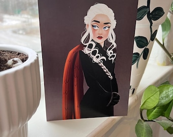 May include: A print of a woman with long white hair wearing a black dress and a red cloak. The woman has blue eyes and a serious expression.