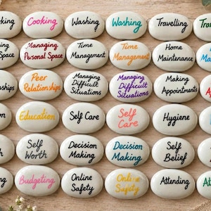 May include: A collection of white, smooth, oval-shaped stones, each featuring a unique word or phrase printed in various colours. The text includes terms like Cooking, Washing, Travelling, Managing Personal Space, Time Management, Home Maintenance, Peer Relationships, Managing Difficult Situations, Making Appointments, Education, Self Care, Hygiene, Self Worth, Decision Making, Self Belief, Budgeting, Online Safety, and Attending.
