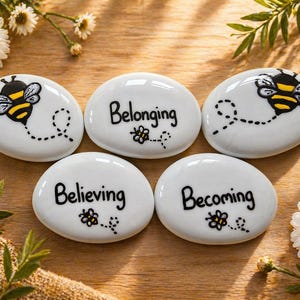 Op de afbeelding: Vijf witte, ovale stenen met zwarte letters en bijenillustraties. De stenen luiden "Belonging", "Believing" en "Becoming". Twee stenen hebben een bijengrafiek. De stenen zijn gerangschikt op een houten oppervlak met bloemaccenten.
