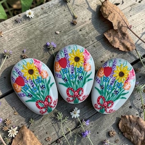 May include: Three painted stones with floral bouquets. Each stone features a bouquet of yellow sunflowers, red tulips, and blue and purple wildflowers tied with a red bow. The stones are on a weathered wooden surface.