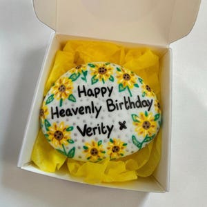 May include: An oval cake decorated with yellow sunflowers and green leaves, with the text "Happy Heavenly Birthday Verity". The cake is presented in a white box with yellow tissue paper. The cake is a birthday gift.