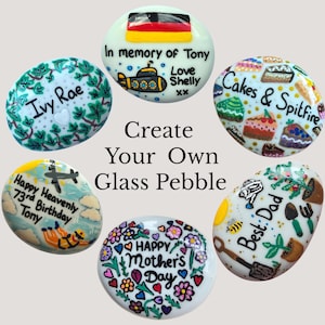 May include: Six smooth, oval glass pebbles featuring hand-painted designs and text. One displays a German flag and submarine, another says Ivy Rae, one features cake illustrations, one says Happy Heavenly 73rd Birthday Tony, one says Happy Mother's Day, and one says Best Dad with gardening tools. The center text reads Create Your Own Glass Pebble.