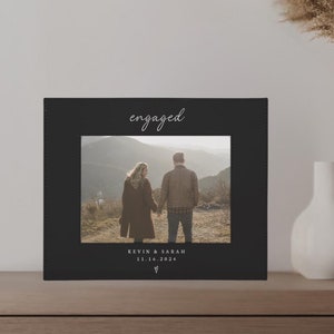 May include: A black and white photo of a couple holding hands and walking away from the camera. The photo is framed with a black border and the word "engaged" is written in white script at the top. The text "Kevin & Sarah 12.18.2022" is written below the photo.