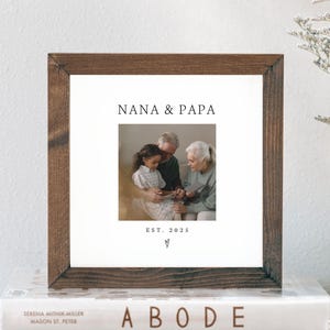 May include: A framed photograph with the text "NANA & PAPA" and "EST. 2025". The photo shows an older couple with a young girl. The frame is dark brown, and the photo is on a stack of books.