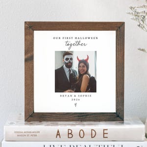 May include: A square wooden frame containing a white print with the text "OUR FIRST HALLOWEEN together" above a photograph of two people in costumes, with the names "BRYAN & SOPHIE" and the year "2026" printed below the image.