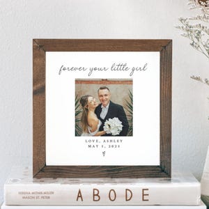 May include: A framed photograph with a dark wood frame, featuring a couple. The text "forever your little girl" is above the photo. Below the photo are the words "LOVE, ASHLEY MAY 5, 2025". The frame rests on a stack of books.