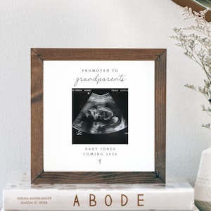May include: A framed ultrasound scan print with the text "PROMOTED TO grandparents" and "BABY JONES COMING 2026." The dark brown frame sits on a stack of books. A white vase with dried flowers is positioned to the right of the frame.