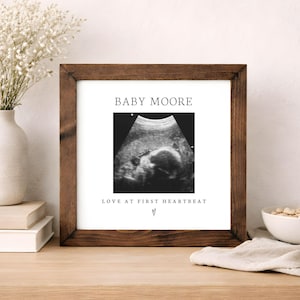 May include: A framed ultrasound print with a dark brown wooden frame. The print displays a grayscale image of a baby. The text "BABY MOORE" is above the image, and "LOVE AT FIRST HEARTBEAT" is below.