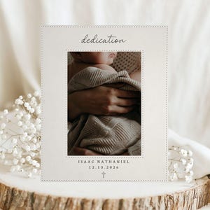 Könnte beinhalten: Eine rechteckige, cremeweiße Widmungskarte mit einem zentralen Foto eines in eine strukturierte Decke eingewickelten Babys. Die Karte enthält den Text dedication, den Namen ISAAC NATHANIEL, das Datum 12.13.2026 und ein kleines Kreuzsymbol.