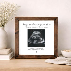 May include: A framed ultrasound scan in a dark brown wooden frame. The image includes the text "hi grandma + grandpa, I can't wait to meet you!" and "BABY ANDERSON DUE DECEMBER 2026". The ultrasound image is in black and white.