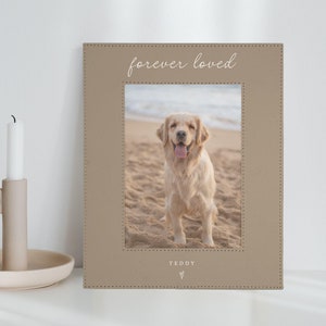 May include: A brown leather photo frame with a stitched border. The frame has a photo of a golden retriever dog on a beach. The text "forever loved" is printed on the top of the frame and the text "TEDDY" is printed on the bottom of the frame with a heart symbol.
