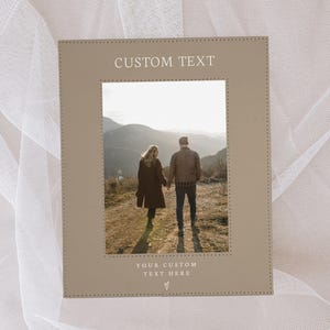 May include: A brown leather picture frame with a stitched border. The frame has a photo of a couple walking in a mountainous landscape. The text "CUSTOM TEXT" is at the top of the frame and "YOUR CUSTOM TEXT HERE" is at the bottom.