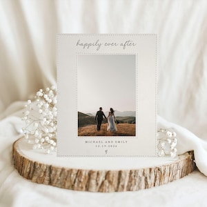 May include: A rectangular off-white card featuring a central photograph of a couple walking in a field. The card has a stitched border design, the text "happily ever after" at the top, and "MICHAEL AND EMILY 12.19.2026" with a small heart icon below.