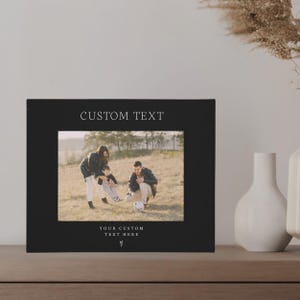 May include: Black photo frame with a white border and a photo of a family of three playing with a soccer ball in a field. The frame has the text "CUSTOM TEXT" at the top and "YOUR CUSTOM TEXT HERE" at the bottom.