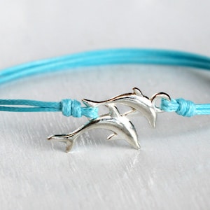 May include: A light blue cord bracelet featuring a silver-toned metal charm shaped like two dolphins swimming together. The cord is secured with knots on either side of the dolphin charm, creating a simple and adjustable design for the wrist.