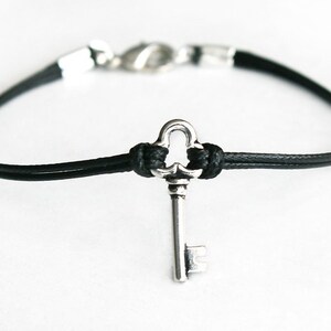 May include: A black leather bracelet with a silver key charm. The key has a decorative, ornate design.