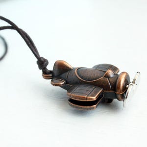 May include: A bronze-colored airplane pendant with a silver propeller hangs from a dark brown cord. The small, detailed plane features a classic design with visible wings, a cockpit, and a round engine.