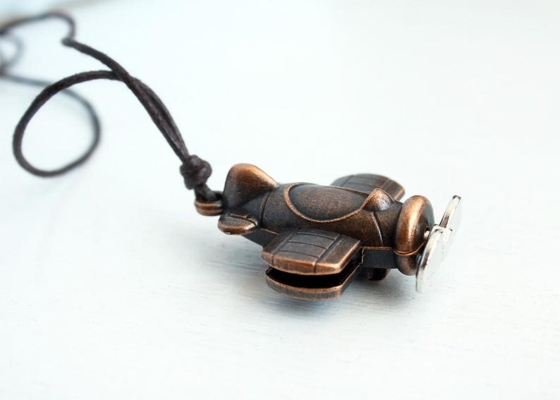 May include: A bronze-colored airplane pendant with a silver propeller hangs from a dark brown cord. The small, detailed plane features a classic design with visible wings, a cockpit, and a round engine.