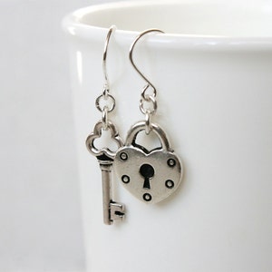 May include: Silver heart-shaped lock and key dangle earrings. The lock has a keyhole and small decorative circles around the edge. The key has a decorative clover-shaped top.